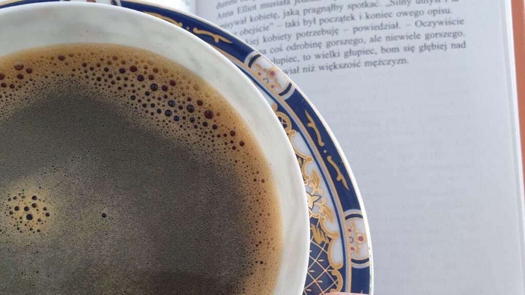 Zbliżenie na filiżankę czarnej kawy w ozdobnej, granatowo-złotej porcelanie, stojącą obok otwartej książki. Zdjęcie symbolizuje analogowy odpoczynek i antidotum na FOMO