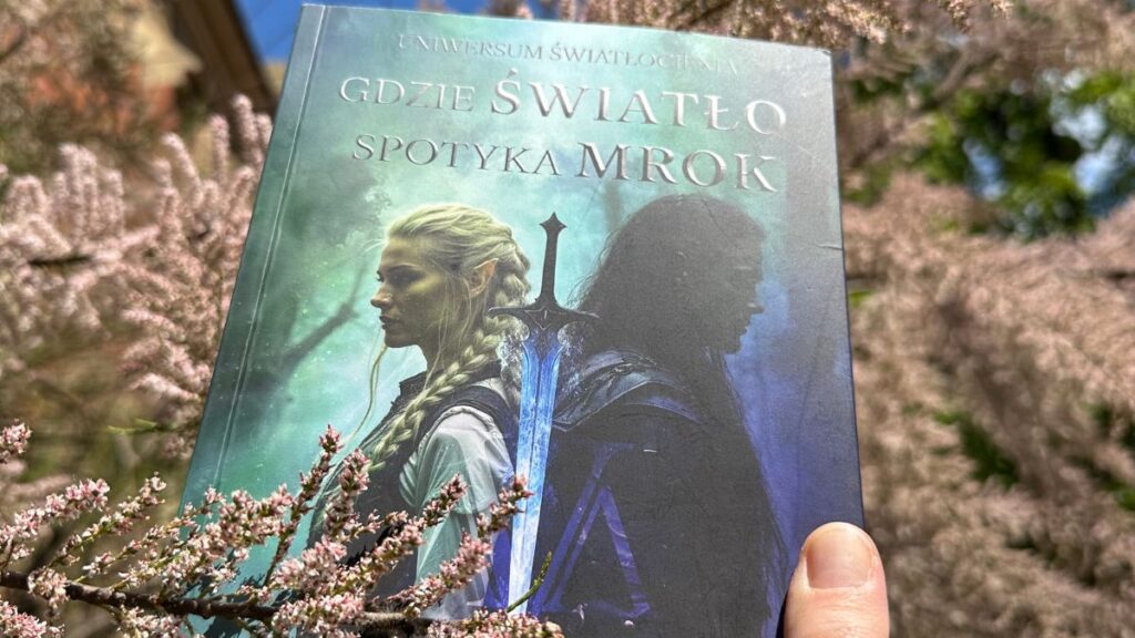 Dłoń trzyma powieść fantasy Leny Klimki pod tytułem „Gdzie Światło spotyka Mrok” na tle kwitnącego na różowo krzewu. Jest to historia, w której kluczową rolę odgrywa natura i magia drzew.