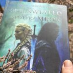 Dłoń trzyma powieść fantasy Leny Klimki pod tytułem „Gdzie Światło spotyka Mrok” na tle kwitnącego na różowo krzewu. Jest to historia, w której kluczową rolę odgrywa natura i magia drzew.